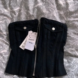 Svart korsett topp - Snygg svart korsett topp i jeansmaterial med dragkedja fram och knappar upptill. Passar bra till både fest och vardag. Storlek S.