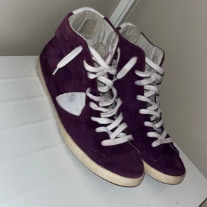 Lila sneakers med vita detaljer - Säljer ett par snygga lila sneakers i mocka med vita snören och detaljer. De har en cool retrostil och är perfekta för vardagsbruk. Skorna har en vit sula och är i bra skick med lite slitage på sulan. Perfekta för dig som vill ha en unik stil! Philippe modell