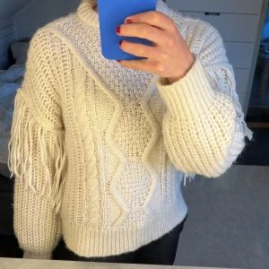 Krämig vit stickad tröja med fransar  - Mysig vit stickad tröja med fransar på ärmarna. Perfekt för kyliga dagar med sitt tjocka material och snygga kabelstickade mönster. Tröjan har en normal passform och långa ärmar, vilket gör den idealisk för både höst och vinter. Ett måste för garderoben!