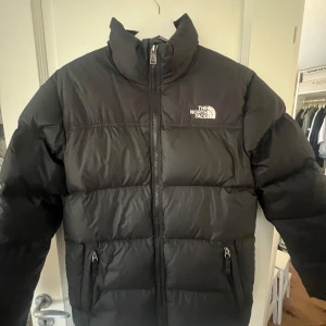 Svart pufferjacka från The North Face - Säljer en svart pufferjacka från The North Face i mycket bra skick. Jackan har en dragkedja framtill och två praktiska fickor med dragkedjor. Perfekt för kalla vinterdagar och håller dig varm och bekväm. Stor logga på bröstet och ryggen. Passar både till vardags och utomhusaktiviteter.