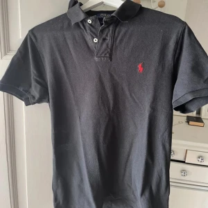 Svart pikétröja från Ralph Lauren - Säljer en klassisk svart pikétröja från Ralph Lauren i storlek M. Tröjan har en röd broderad logga på bröstet och är i slim fit. Perfekt för både vardag och fest! 🖤