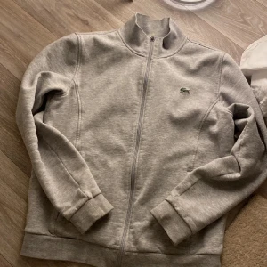 Grå Lacoste zip-tröja  - Säljer e  grå zip-tröja från Lacoste i mycket bra skick. Använd fåtal gånger. Den har en dragkedja framtill och det klassiska krokodilmärket på bröstet. Perfekt för både vardag och träning. Passar bra till till alla årstider. Nypris ös 1200