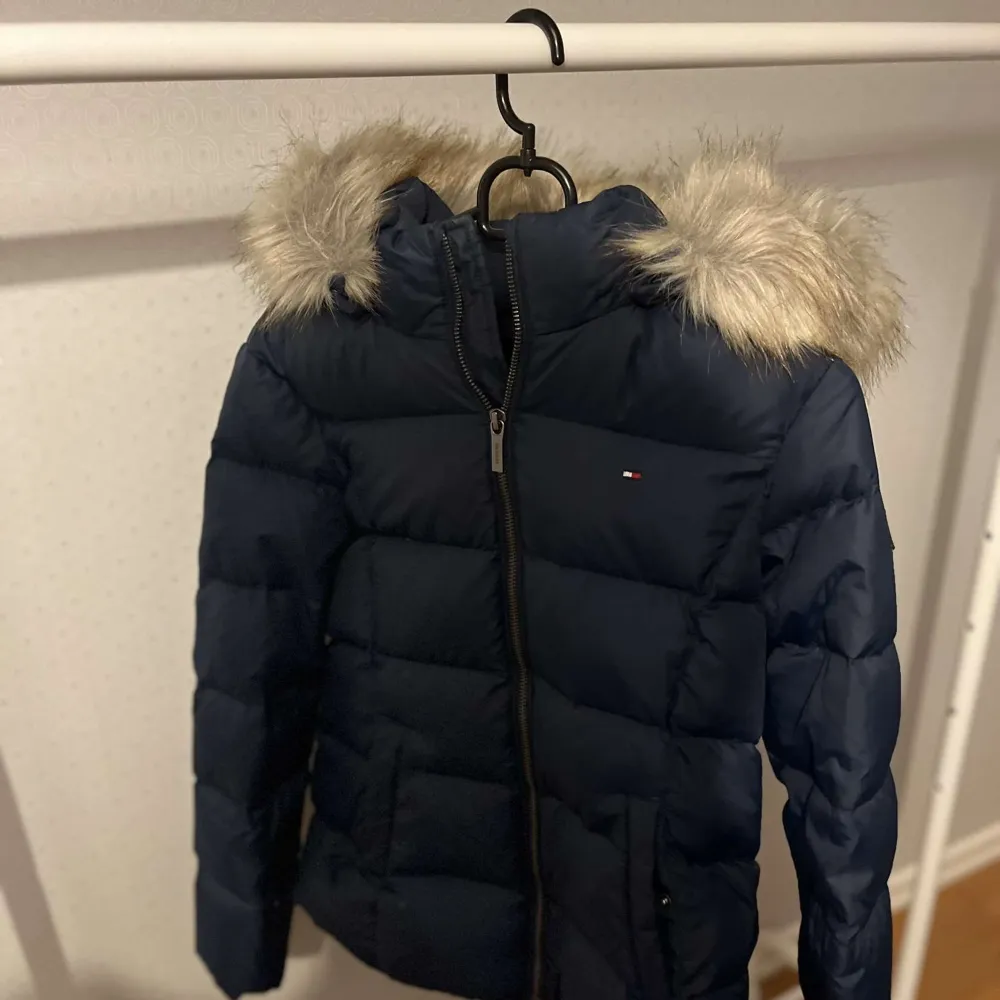 Säljer en snygg mörkblå dunjacka från Tommy Hilfiger. Jackan har en mysig pälskantad huva och dragkedja framtill. Perfekt för kalla vinterdagar! Den är i mycket bra skick och redo för nya äventyr. 🧥❄️. Jackor.