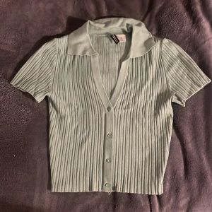 Grön ribbad topp från H&M - Säljer en snygg grön ribbad topp från H&M i storlek XXS. Den har korta ärmar och knappar framtill, perfekt för både vardag och fest. Toppen är i mycket bra skick och har en stilren krage. Passar perfekt till både jeans och kjol!