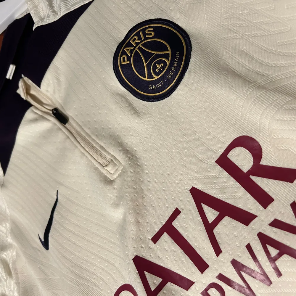 Säljer en snygg beige fotbollströja från Nike med PSG-logga och Qatar Airways sponsortryck. Tröjan har korta ärmar och en stilren design med mörka detaljer vid kragen. Perfekt för fotbollsfans som vill ha något unikt i garderoben! 🏆byxa går att Få med . T-shirts.