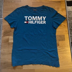 Blå t-shirt från Tommy Hilfiger - Säljer en snygg blå t-shirt från Tommy Hilfiger i ekologisk bomull. Den har en klassisk passform med korta ärmar och det ikoniska Tommy Hilfiger-logotypen tryckt på framsidan. Perfekt för en avslappnad stil eller vardagsbruk.      Använd ett fåtal gånger.  Strl 176 (sitter som en S)