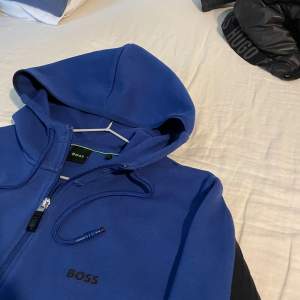 Säljer en snygg blå BOSS hoodie med svarta detaljer, Storlek S men passar M. Köpt på Hennings 1799kr. Perfekt för både vardag och träning. Hoodien är ny och använd fåtal gångar.