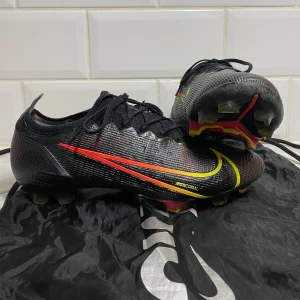 Nike Mercurial fotbollsskor - Säljer ett par grymma Nike Mercurial fotbollsskor i svart med coola röda och gula detaljer. Skorna har en snygg och modern design med snörning för perfekt passform. Perfekta för fotbollsspelare som vill ha både stil och funktion på planen. Använda men i bra skick! Info: Denna sko är utrustad med FG-dobbar och ska enbart användas på naturgräs. Vikt: 205 gram Skick: 8,5/10 Använda: vid 10-20 tillfällen  Inga synliga skador OBS! förutom sulan har ett märke från skohornet.