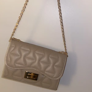 Beige axelväska med guldkedja - Snygg beige axelväska i quiltat mönster med en elegant guldkedja som axelrem. Perfekt för både vardag och festliga tillfällen. Väskan har en stilren design och är i mycket bra skick.
