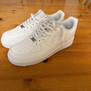 Vita Nike Air Force 1 sneakers - Säljer ett par klassiska vita Nike Air Force 1 i nytt skick. De är helt vita med snörning och har den ikoniska Nike-loggan på sidan och hälen. Perfekta för både vardag och fest! 