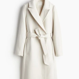 Vit kappa med bälte - Säljer en elegant vit kappa i mycket bra skick. Används endast 4-5 gånger. Den har en klassisk design med långa ärmar och ett bälte i midjan för en snygg passform. Perfekt för höst och vår när du vill se stilren ut. Passar både till vardags och festliga tillfällen.