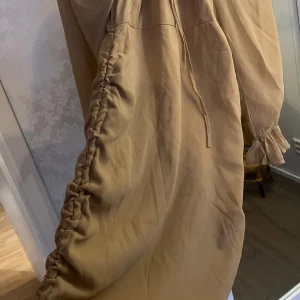 Beige klänning med volangdetaljer - Säljer en superfin beige klänning med volangdetaljer längs sidorna. Den har långa ärmar med resår och volang vid ärmsluten. Perfekt för höst och vår! Klänningen har en knytning framtill som ger en söt touch. Passar både till vardags och festligare tillfällen.