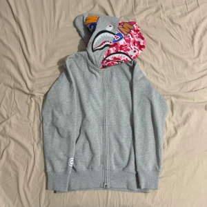Grå BAPE hoodie med rosa camoluva - Säljer en grå hoodie från BAPE med det ikoniska hajmotivet på luvan. Den har en dragkedja framtill och färgglada bokstäver 'WGM' på luvan. Luvan i insidan har ett coolt rött och rosa kamouflagemönster. Perfekt för streetwear-stilen! Det går även att stänga till hoodien ända upp till luvan så det blir en mask. Passar som M-L