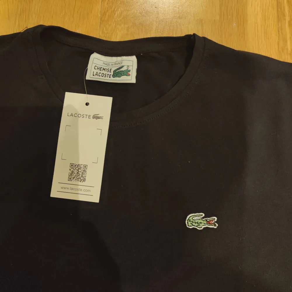 Säljer en klassisk svart t-shirt från Lacoste i nytt skick. Den har en normal passform och är kortärmad, perfekt för vardagsbruk. T-shirten är gjord av bomull och har den ikoniska krokodilloggan på bröstet. Ett måste för garderoben! 🐊. T-shirts.