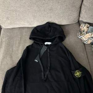 Säljer en snygg svart hoodie från Stone Island. Den har en klassisk design med huva och en ikonisk logga på ärmen. Perfekt för en casual look eller att bara mysa i. Helt ny med taggar kvar!