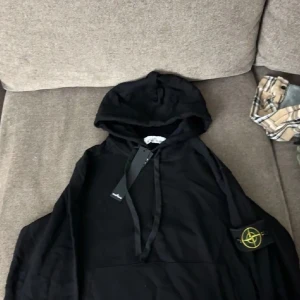 Svart hoddie från Stone island - Säljer en snygg svart hoodie från Stone Island. Den har en klassisk design med huva och en ikonisk logga på ärmen. Perfekt för en casual look eller att bara mysa i. Helt ny med taggar kvar!