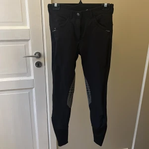 Svarta ridbyxor från CRW - Säljer ett par svarta ridbyxor från CRW i storlek 38. Byxorna har snygga detaljer med dragkedjor och små nitar. Perfekta för ridning med en tight passform som ger bra stöd. De är i mycket bra skick och redo för nya äventyr i stallet!
