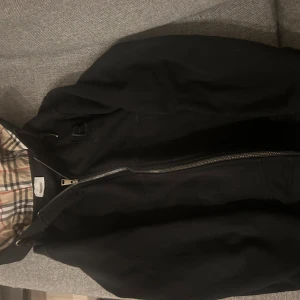 Svart hoodie från Burberry - Säljer en snygg svart jacka från Burberry i bra skick. Den har en klassisk rutig insida och dragkedja framtill. Perfekt för höst och vår! Jackan har justerbara snören vid huvan för extra komfort. Passar både till vardags och lite finare tillfällen. Har inte använd hoodien så mycket den är i bra skick❤️