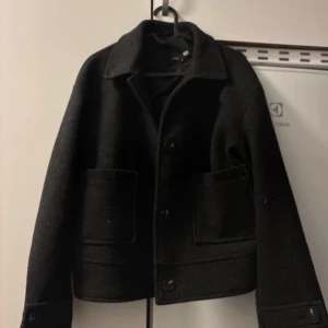 Svart kappa från 157 - Säljer en stilren svart kappa från 157 i storlek M. Den har en klassisk design med knappar framtill och stora fickor. Perfekt för höst och vinter, och ger en elegant touch till vilken outfit som helst. Jackan är i mycket bra skick och redo för nya äventyr!