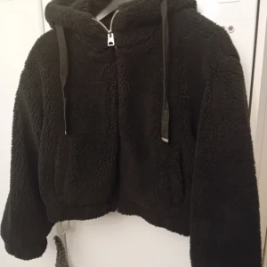Svart teddyjacka med huva - Mysig svart teddyjacka med huva och dragkedja framtill. Perfekt för kyliga dagar när du vill vara både varm och stilren. Jackan har långa ärmar och en skön, fluffig känsla. Passar bra till både vardag och avslappnade tillfällen.