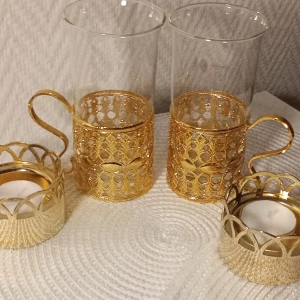 Guldfärgade värmeljushållare och glas med hållare - Säljer ett set med två eleganta glas med guldfärgade hållare och matchande ljushållare. De är till för Irish coffee men kan användas till all form av dryck. Glasen har en vacker, detaljerad design med handtag, perfekt för att ge en lyxig känsla till ditt hem. Ljushållarna har ett liknande mönster och passar perfekt för värmeljus. Perfekt för att skapa en mysig atmosfär!