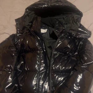 Svart pufferjacka från Calvin Klein - Säljer en snygg svart pufferjacka från Calvin Klein. Den är i glansigt material och har en dragkedja framtill. Perfekt för kalla dagar med sin varma och vadderade design. Jackan har en huva och CK-loggan på bröstet. Passar perfekt för vintern!