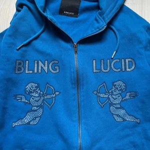 Blå hoodie från Bling Lucid - Säljer en cool blå hoodie från Bling Lucid med dragkedja och blingade detaljer. Den har texten 'BLING LUCID' och två änglar i strass på framsidan. Perfekt för att sticka ut i mängden! 💎