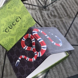 Svart Gucci Plånbok - Säljer en snygg svart plånbok från Gucci med ett coolt ormmotiv i rött, vitt och blått. Plånboken har flera kortfack och en transparent ficka för ID-kort. Den kommer i en elegant grön förpackning med Gucci-loggan. Perfekt för att ge din stil en lyxig touch!