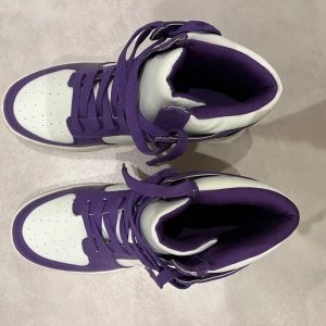 Lila och vita sneakers - Säljer ett par snygga lila och vita sneakers i mycket bra skick. De har en cool design med snörning och hög krage för extra stöd. Perfekta för både vardag och festliga tillfällen. Sulan är vit och i bra skick, vilket ger en fräsch look.
