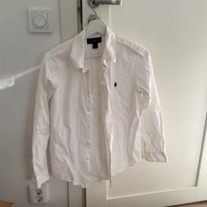 Vit skjorta från Ralph Lauren - Säljer en klassisk vit skjorta från Ralph Lauren i storlek 18. Skjortan är i bomull och har långa ärmar med knappar framtill. Perfekt för både vardag och festliga tillfällen. Den har en broderad logga på bröstet som ger en stilren touch. Barnstorlek 18