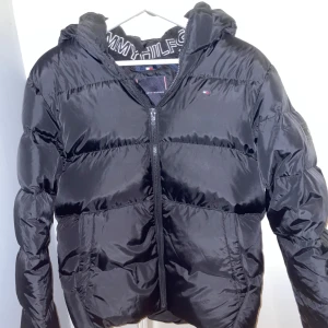 Svart vinterjacka från Tommy Hilfiger XS - Säljer en svart jacka från Tommy Hilfiger i storlek 164cm men sitter bra på mig som är 170. Jackan är i nytt skick. Perfekt för kalla vinterdagar. Den har en luva för extra värme. Jackan har ett Tommy Hilfiger märke på bröstet och även på vänster arm. Jackan är köpt för 2000kr och har endast provats hemma. Bilderna blev ganska ljusa och jackan såg lite glansig ut men jackan är helt svart. Jag säljer den på grund av att den var lite för stor. Hör gärna av er vid frågor.  Priset går att diskutera.
