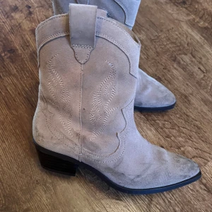 Beige cowboy boots i mocka - Säljer ett par snygga beige cowboy boots i mocka. De har en spetsig tå och låg klack, perfekt för en avslappnad men stilren look. Bootsens detaljerade sömmar ger dem en unik touch. Passar perfekt till höst- och våroutfits!