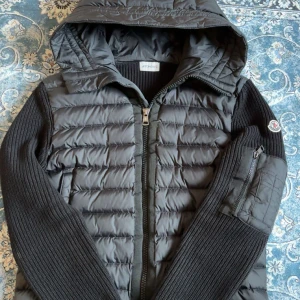 Svart Moncler cardigan - Säljer en snygg svart cardigan jacka från Moncler. Den har en huva och dragkedja framtill. Ärmarna är stickade och har en praktisk ficka med dragkedja. Perfekt för höst och vinter, håller dig varm och stilren. Passar både till vardags och lite finare tillfällen. NFC och Scan fungerar utan problem. Knappast använd inga tecken på användning. 