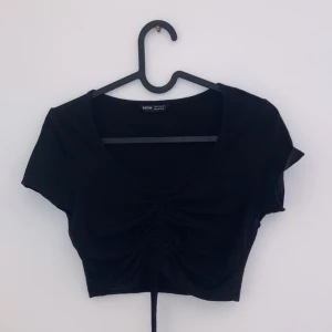 Svart croppad topp från Shein - Snygg svart croppad topp från Shein med korta ärmar och en knytning framtill som ger en fin rynkad effekt. Perfekt för sommarens alla fester eller en avslappnad dag ute. Toppen är i ett mjukt och stretchigt material för bästa komfort.