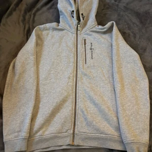 Grå huvtröja från Sail Racing - Säljer en grå huvtröja från Sail Racing i bra skick. Den har en dragkedja framtill och en broderad logga på huvan. Perfekt för höst och vår, med långa ärmar och mjukt material för extra komfort. Passar både till vardags och lättare aktiviteter.