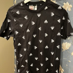 Svart t-shirt med mönster från Adidas - Säljer en svart t-shirt från Adidas i bra skick. Den har ett coolt mönster med vita Adidas-loggor över hela tyget. Perfekt för vardagsbruk eller träning. Kortärmad och gjord i mjukt bomullsmaterial för extra komfort.