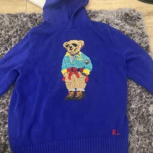 Blå hoodie med björnmotiv från Polo Ralph Lauren - Säljer en supercool blå hoodie från Polo Ralph Lauren med ett broderat björnmotiv på framsidan. Perfekt för att hålla sig varm och stilren under kyliga dagar. Den har en klassisk passform och är gjord i ett mjukt material. Passar perfekt för både vardag och avslappnade tillfällen. Resell pris ligger på 3500.