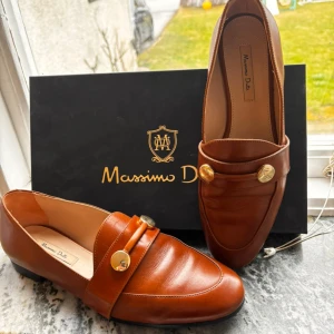 Bruna loafers från Massimo Dutti - Snygga bruna loafers  från Massimo Dutti med dekorativa guldfärgade knappar. Perfekta för en stilren look både till vardags och fest. Skorna har en klassisk design med en bekväm passform.