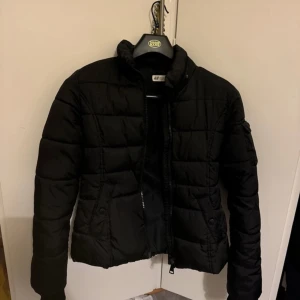 Svart dunjacka från H&M - Säljer en svart dunjacka från H&M i bra skick. Jackan är quiltad och har en dragkedja framtill. Perfekt för höst och vinter med sin varma och bekväma design. Den har långa ärmar och en hög krage för extra skydd mot kylan. Saknar luva