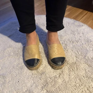 Chanel espadrillos i lammskinn - Säljer ett par lyxiga espadrillos från Chanel i beige och svart lammskinn. De har en snygg Chanel logga på ovansidan och en klassisk flätad sula. Perfekta för en stilren och bekväm look. Använda men i bra skick. Passar perfekt till både vardag och fest. Köpta på Bangkok flygplats ord pris 10480kr idag!