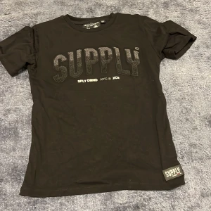 Svart t-shirt från Supply & Demand - Säljer en svart t-shirt från Supply & Demand med texten 'SUPPLY' i stort tryck på framsidan. Den är i bra skick och perfekt för vardagsbruk. Tillverkad i Turkiet och har en normal passform. Passar perfekt till jeans eller shorts för en avslappnad stil.