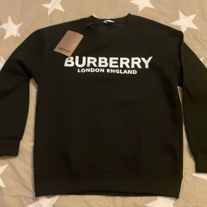 Burberry Crewneck - Säljer en snygg svart sweatshirt från Burberry med vit text 'BURBERRY LONDON ENGLAND' på bröstet. Tröjan är i nytt skick och perfekt för både vardag och fest. Den har en klassisk passform och är superbekväm. Passar perfekt till jeans eller kjol för en stilren look.