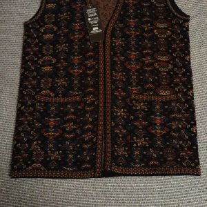 Mönstrad väst med broderi - Säljer en unik och färgglad väst med ett detaljerat broderat mönster. Västen har en v-ringad design och två praktiska fickor framtill. Perfekt för att ge din outfit en bohemisk touch. Passar bra till både vardag och fest!