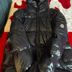 Svart pufferjacka från Moncler - Säljer en snygg svart pufferjacka från Moncler i mycket bra skick. Jackan har en glansig yta med dragkedja och en broderad Moncler-logga på ärmen. Perfekt för kalla vinterdagar med sin varma och stiliga design. Passar både till vardags och festliga tillfällen.