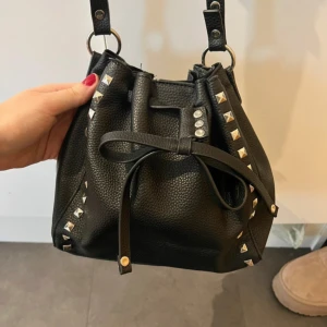 Svart axelväska med nitar - Snygg svart axelväska i fejkskinn med coola nitar längs kanterna. Väskan har en justerbar axelrem och stängs med dragsko. 