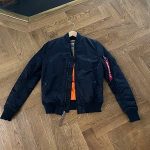Marinblå bomberjacka från Alpha Industries - Säljer en snygg marinblå bomberjacka från Alpha Industries i storlek XS för endast 449 kr. Jackan har en cool orange insida och en dragkedja framtill. Perfekt för höst och vår, med ribbade muddar och en klassisk bomberstil. Den är i mycket bra skick och redo för nya äventyr! 