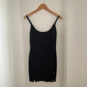 Svart klänning från Zorannah for NA-KD - Snygg svart klänning från Zorannah for NA-KD i storlek XS. Den är tight och kort med snörningsdetaljer på sidorna som ger en edgy look. Perfekt för fest eller en utekväll. Klänningen är ärmlös och har en stilren design.