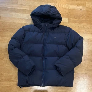 Mörkblå pufferjacka från GANT - Säljer en snygg mörkblå pufferjacka från GANT i storlek 176 cm, perfekt för kalla dagar. Jackan har en praktisk dragkedja och en mysig huva. Den är i mycket bra skick, i princip som ny, inga skador eller skavanker. Passar perfekt för både vardag och utflykter! 💙