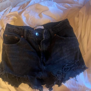 Svarta jeansshorts med fransar - Säljer ett par coola svarta jeansshorts med fransiga kanter. De har en klassisk femficksdesign och en knapp med dragkedja framtill. Perfekta för sommaren och ger en avslappnad look. Passar bra till både vardag och fest!