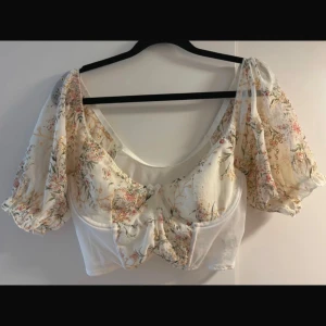 Blommig blus med puffärmar - Säljer en superfin blommig blus med puffiga ärmar. Den är vit med ett romantiskt blommönster i mjuka färger. Perfekt för sommarens fester eller en mysig vårdag. Blusen har en söt volangdetalj framtill och är i mycket bra skick.